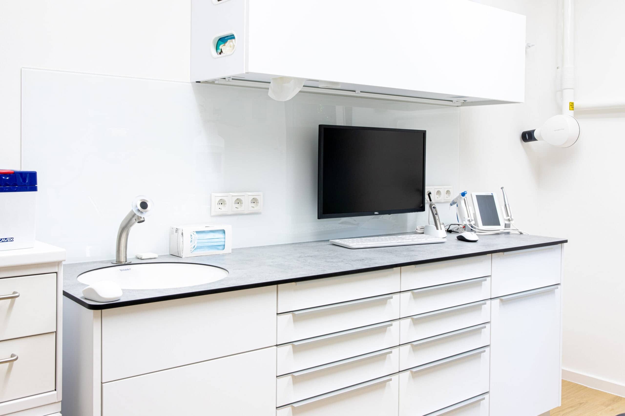 Arbeitszeile im Behandlungszimmer: graue Platte, weiße Schubladenfronten, Wandmonitor und Röntgenstrahler